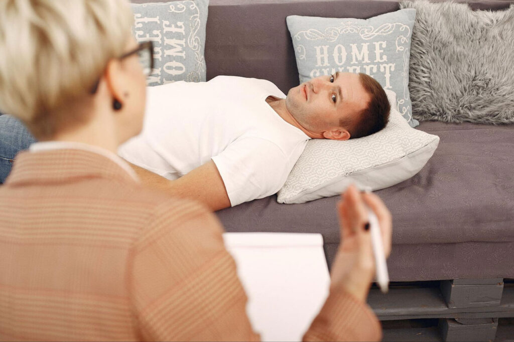 A man lying on a couch speaking with a therapist who is taking notes, representing a safe, supportive environment for processing trauma and working through recovery.