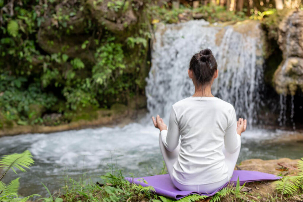 A woman in white athletic wear sits on a purple yoga mat in a meditative pose, viewed from behind, facing a lush green waterfall in a serene forest setting.