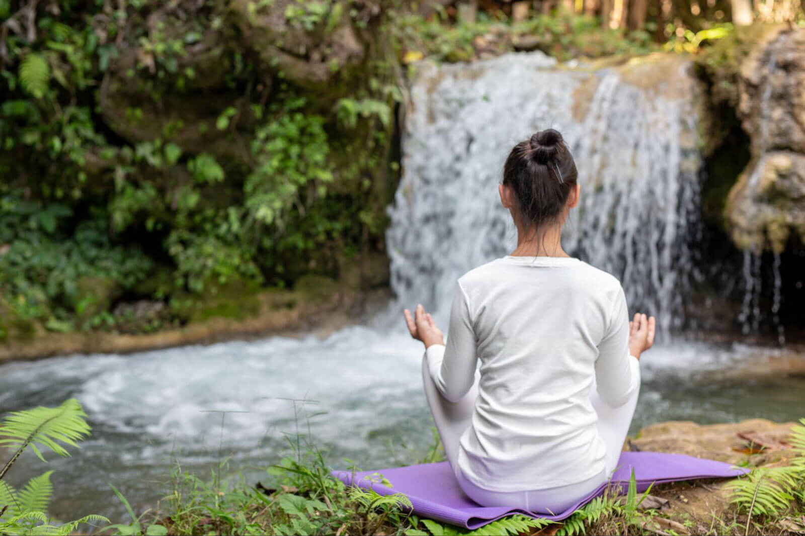 A woman in white athletic wear sits on a purple yoga mat in a meditative pose, viewed from behind, facing a lush green waterfall in a serene forest setting.