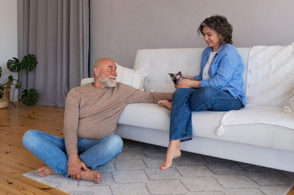 A smiling older man sits cross-legged on the floor while a younger woman on a white sofa holds a small cat, sharing a relaxed and cheerful moment together in a bright living room.