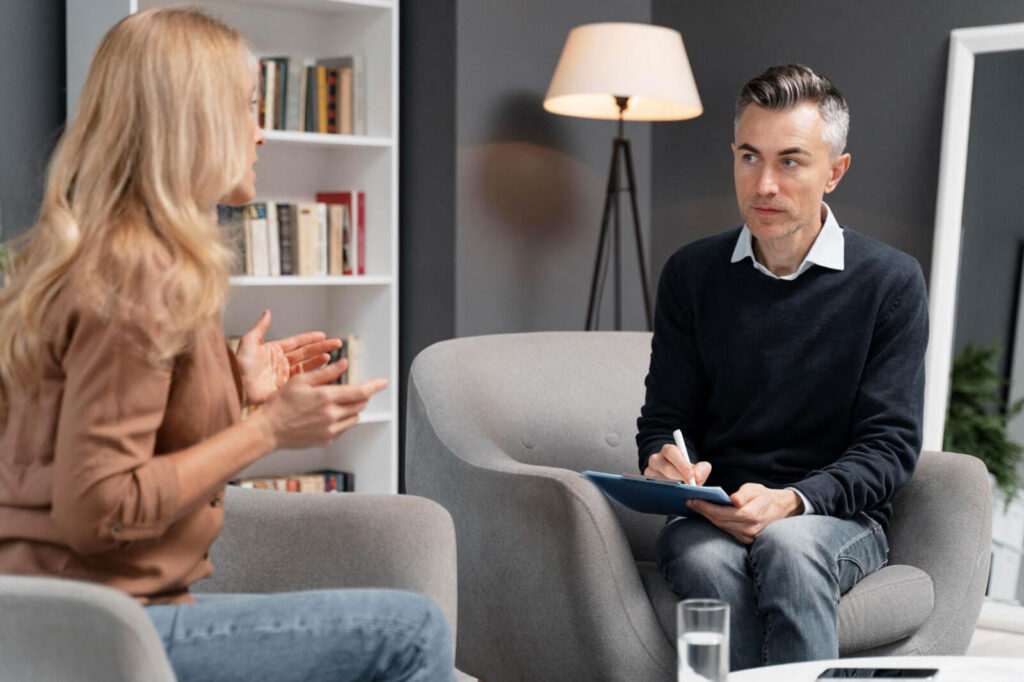 A woman with long blonde hair gestures expressively while speaking to a focused male therapist holding a clipboard, seated across from each other in a dimly lit, modern therapy office.