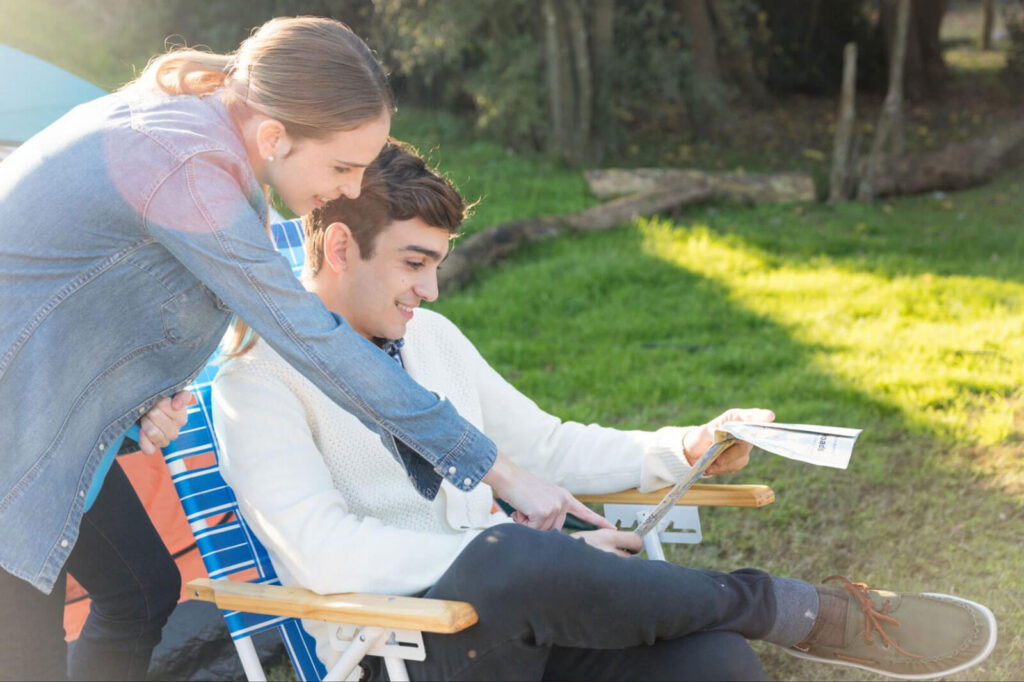 A young woman leans over the shoulder of a smiling young man seated in an outdoor chair, both looking down at a document he's holding in a sunny, grassy setting.