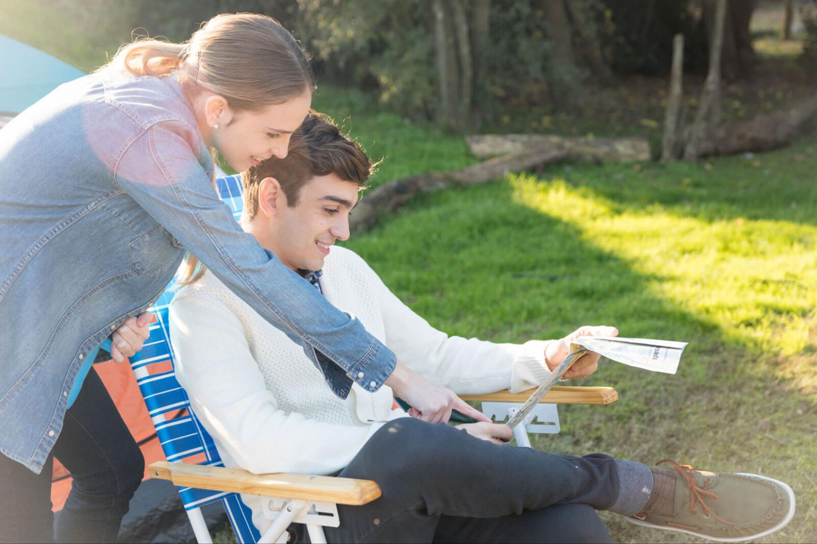 A young woman leans over the shoulder of a smiling young man seated in an outdoor chair, both looking down at a document he's holding in a sunny, grassy setting.