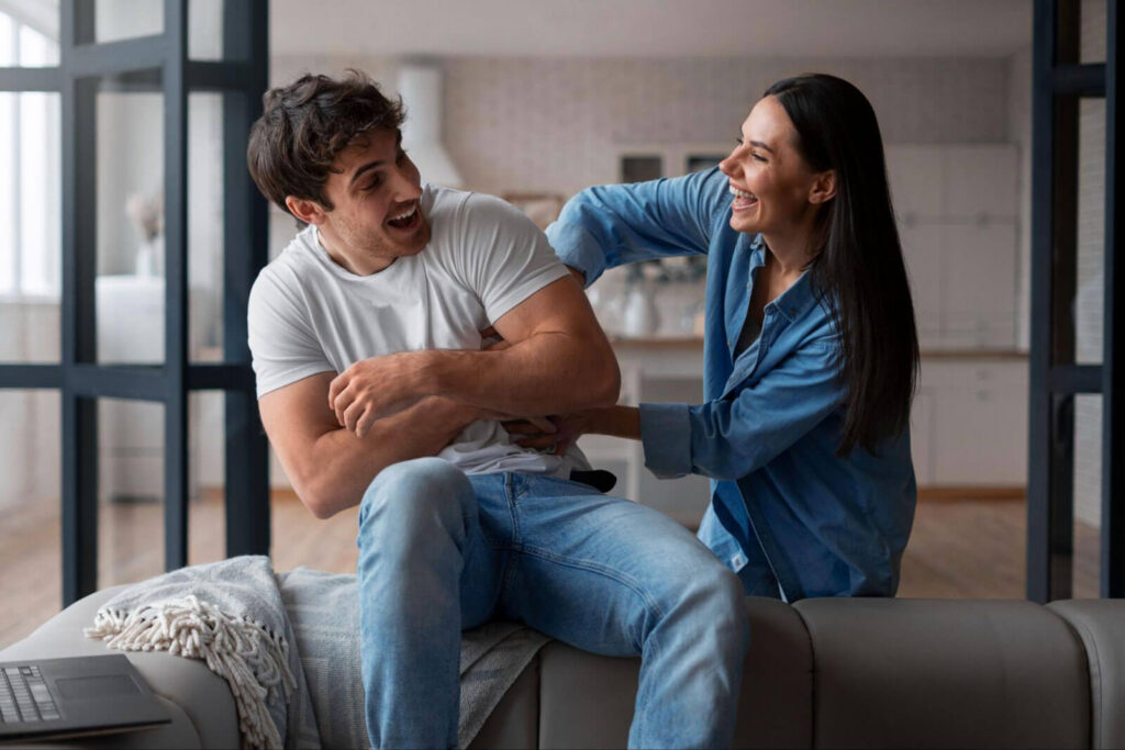 A man and woman laughing together on a couch at home, sitting close in a playful moment.