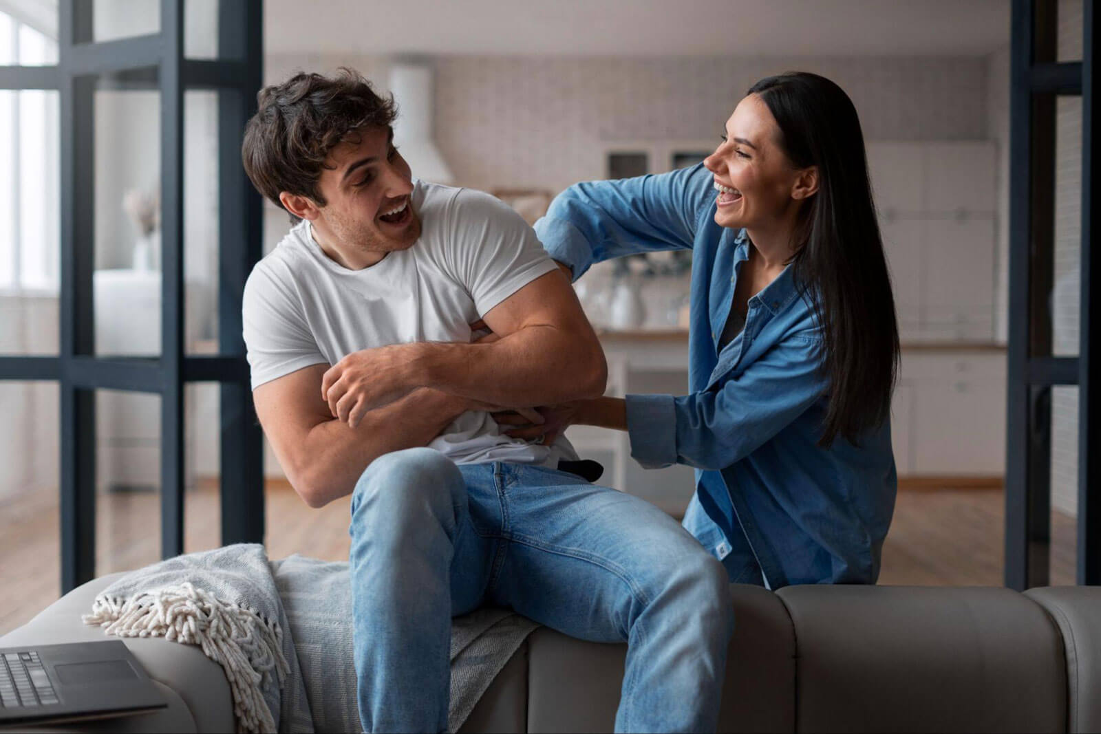 A man and woman laughing together on a couch at home, sitting close in a playful moment.