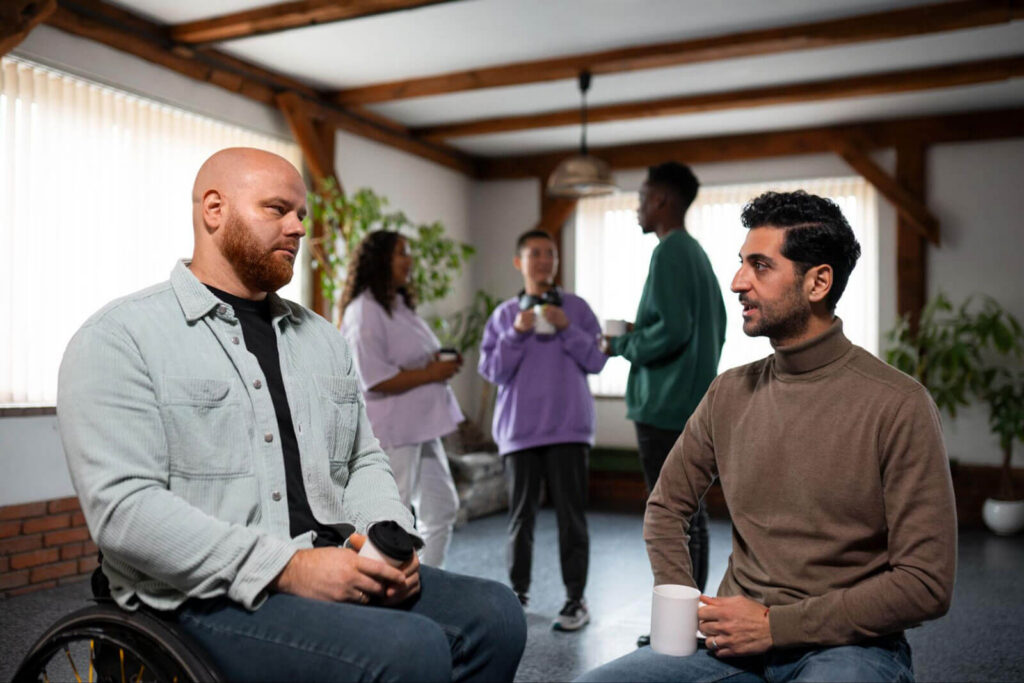 A man in a wheelchair talking with another man holding a coffee mug, with a small group chatting in the background of a sunlit room.