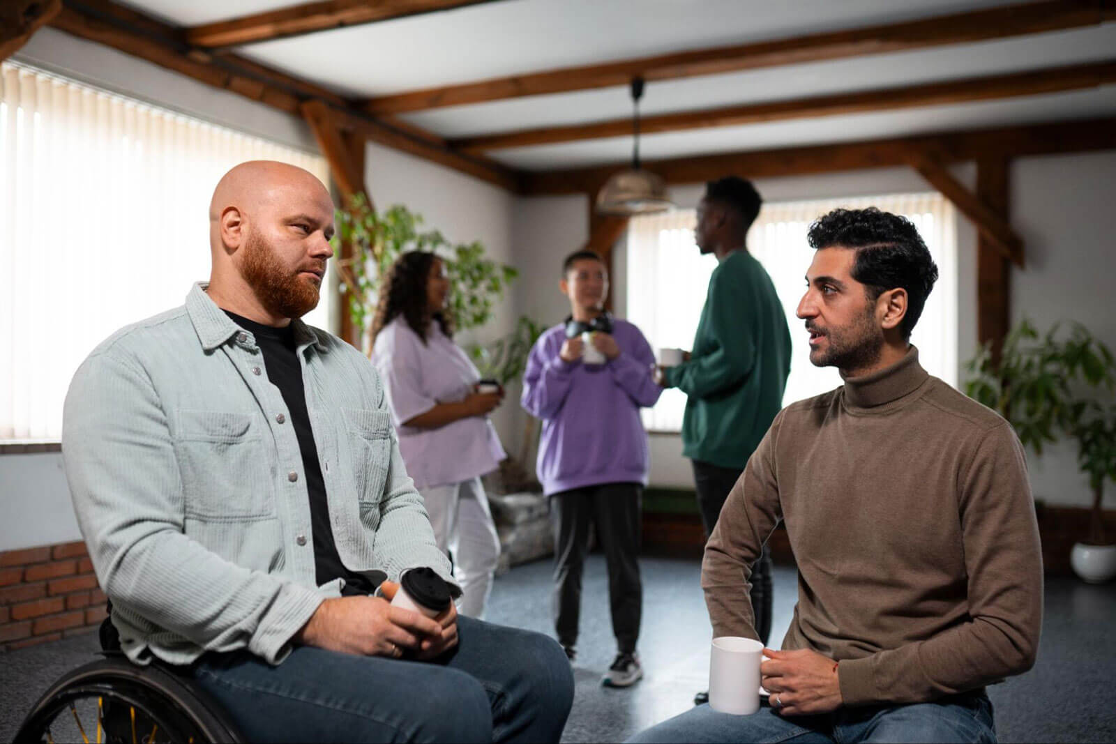 A man in a wheelchair talking with another man holding a coffee mug, with a small group chatting in the background of a sunlit room.