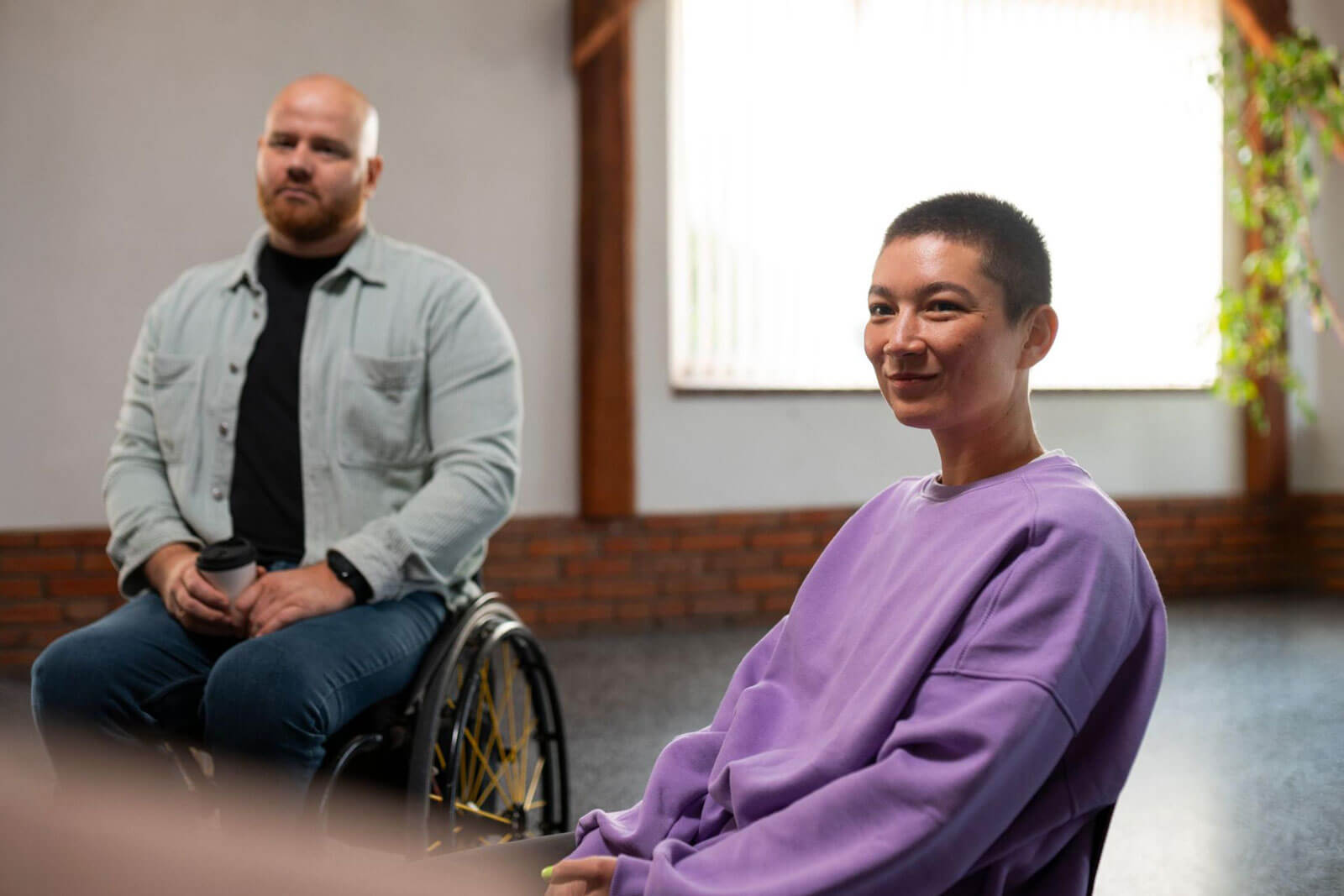 A woman with short hair smiling at the camera while seated, with a man in a wheelchair sitting nearby in a sunlit room.