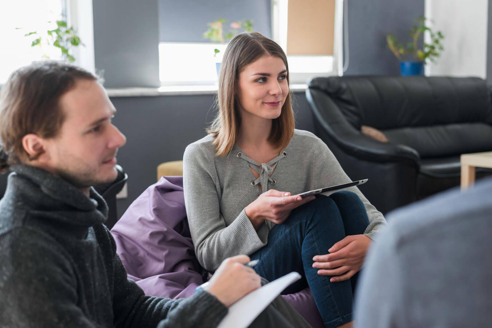 A woman holding a tablet and a man taking notes listen during a group discussion.