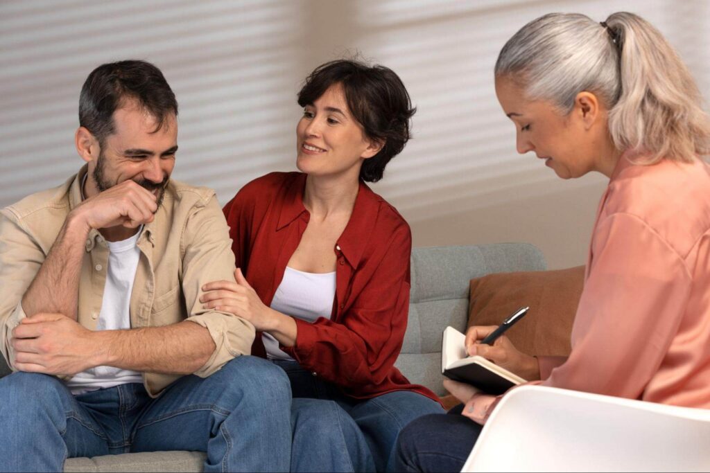 Man laughing while a woman comforts his arm and a counselor takes notes beside them.