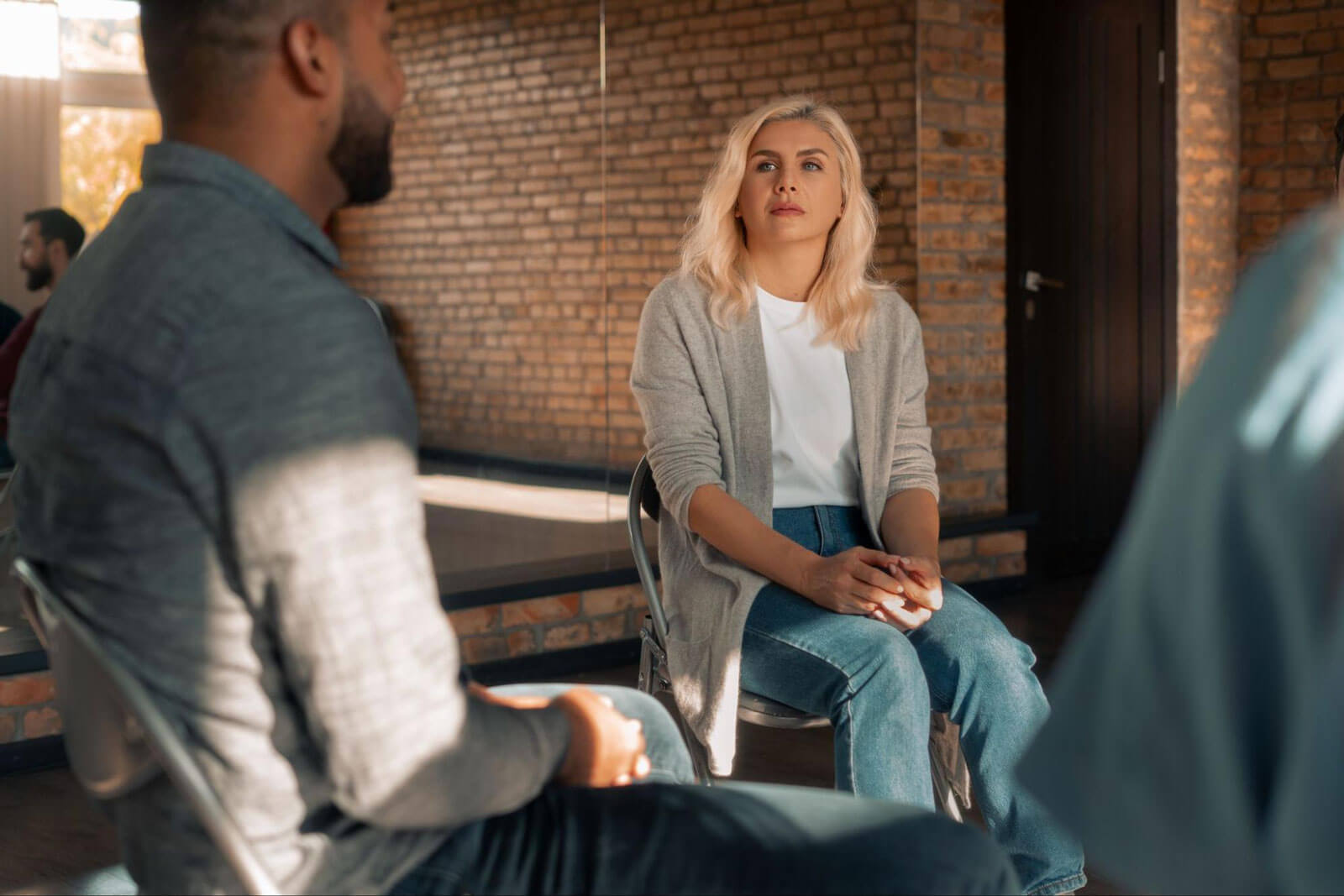 A woman sits with her hands clasped, listening to a man during a group discussion.