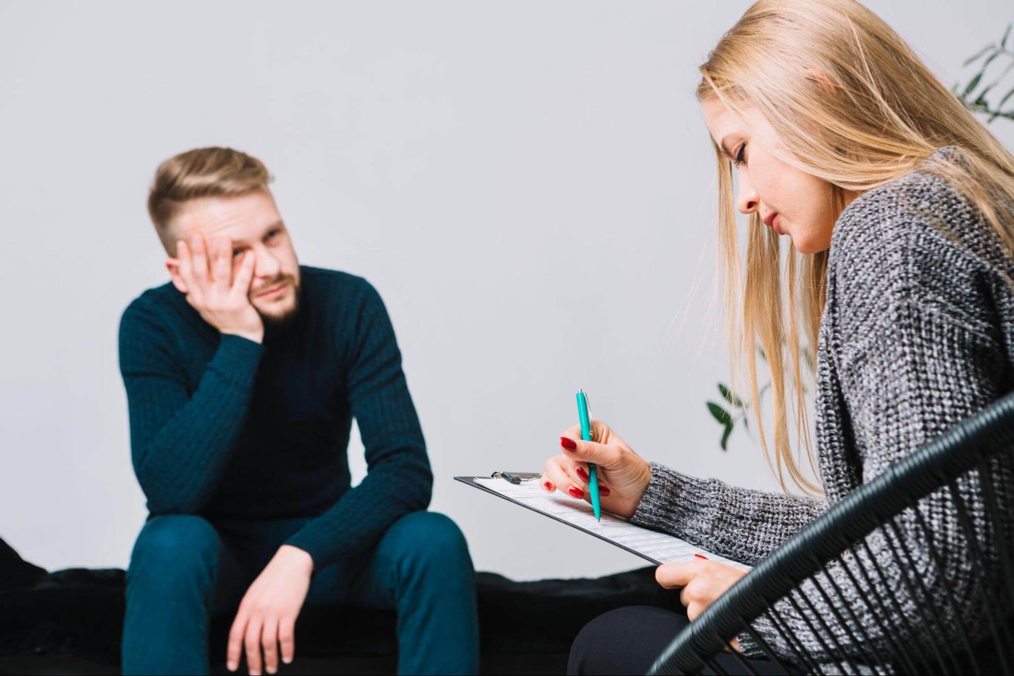 Distressed man covering his face while a woman takes notes on a clipboard.