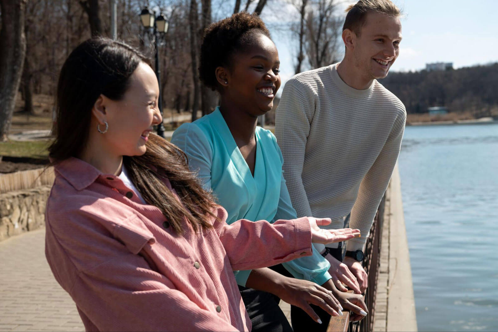 Three people smile and talk while standing beside a lakeside railing.