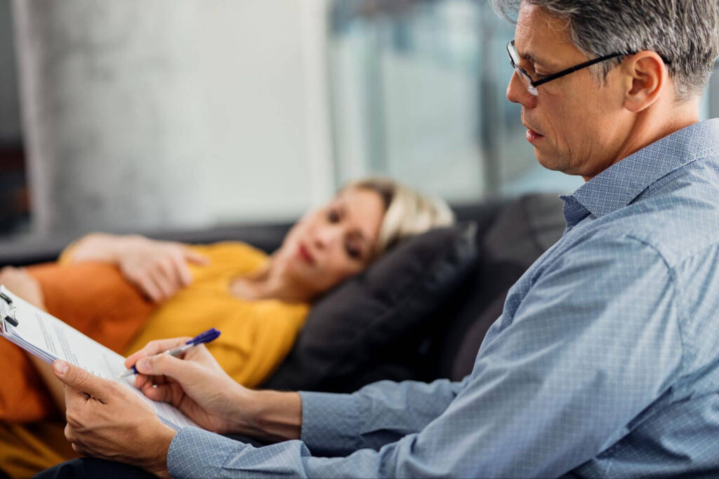 A man takes notes on a clipboard while a woman reclining on a couch speaks with him.