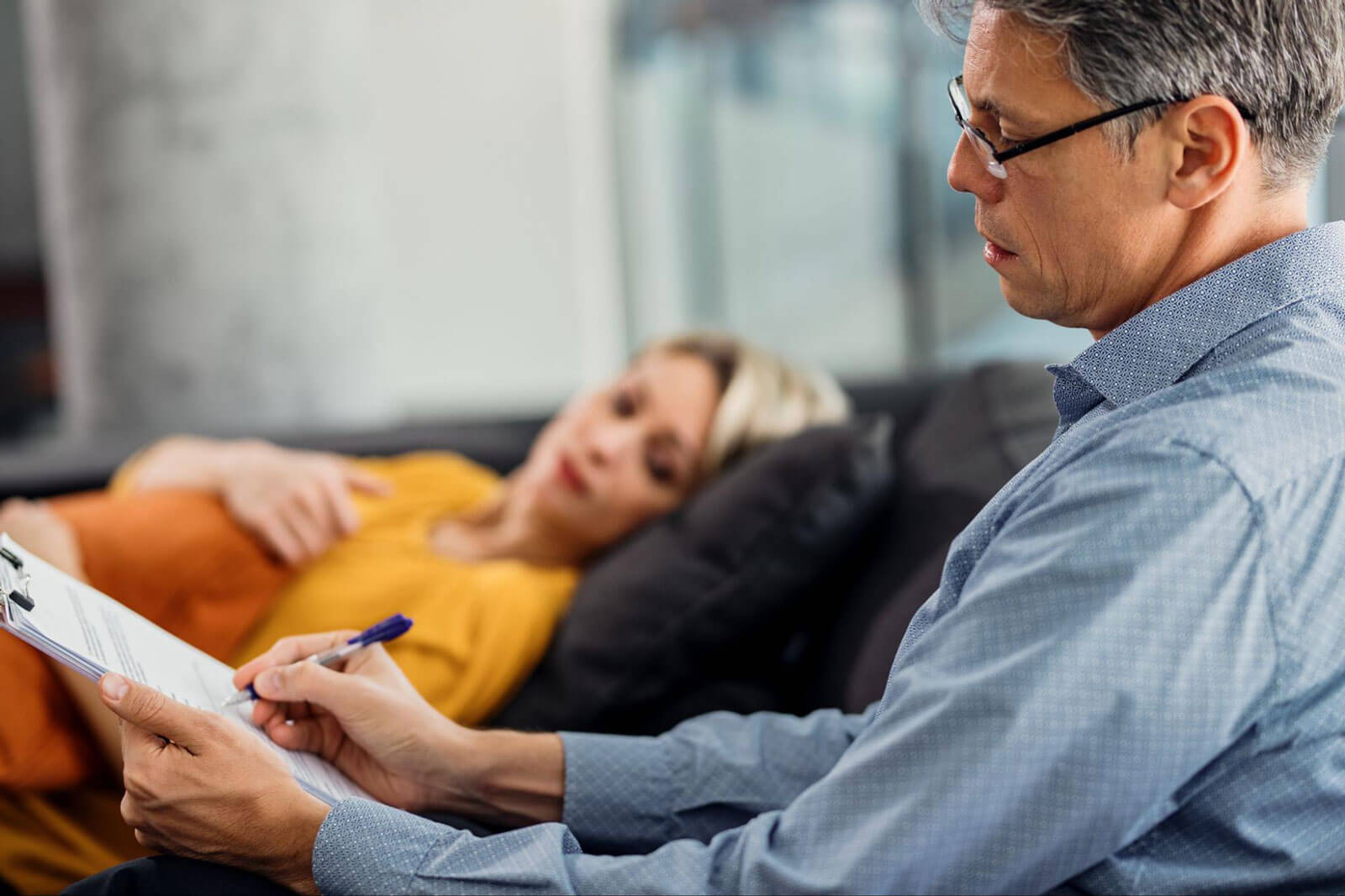 A man takes notes on a clipboard while a woman reclining on a couch speaks with him.