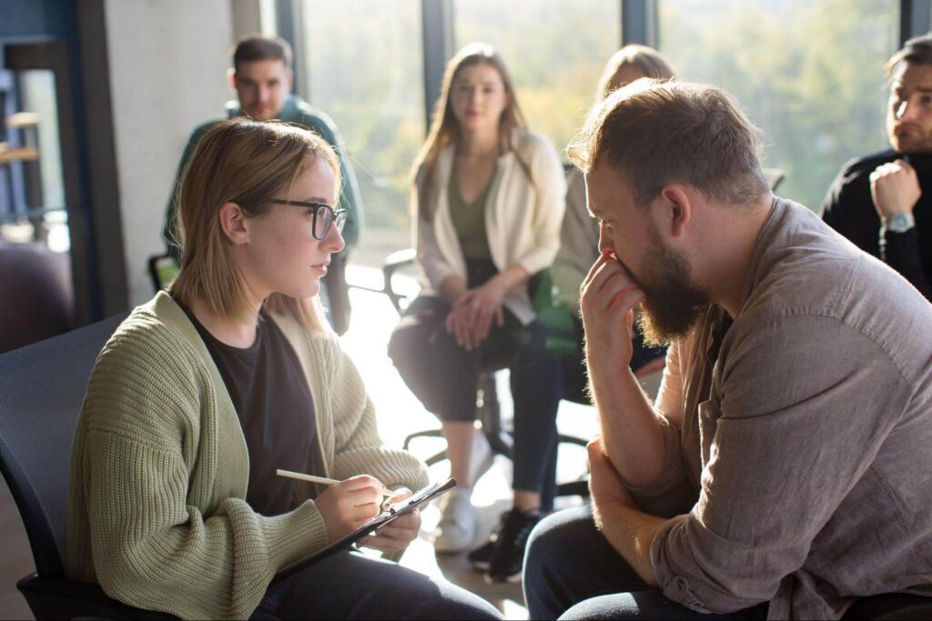 Woman writing on a clipboard while speaking closely with a thoughtful bearded man.