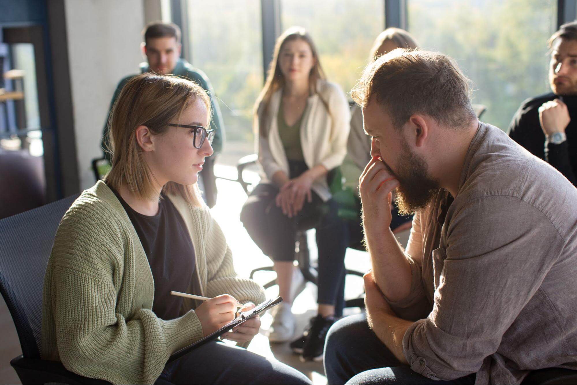 Woman writing on a clipboard while speaking closely with a thoughtful bearded man.